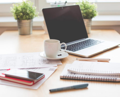 Ein Arbeitsplatz mit Laptop, Handy, Notizbüchern, Stift und Kaffeetasse.