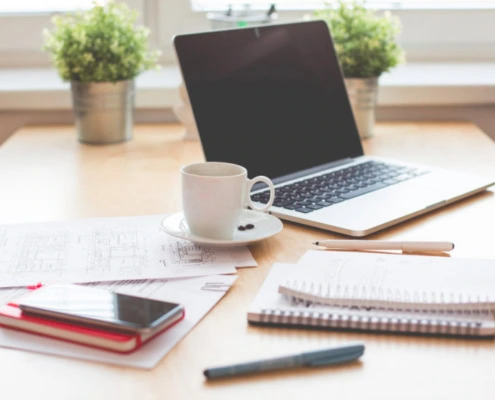 Ein Arbeitsplatz mit Laptop, Handy, Notizbüchern, Stift und Kaffeetasse.