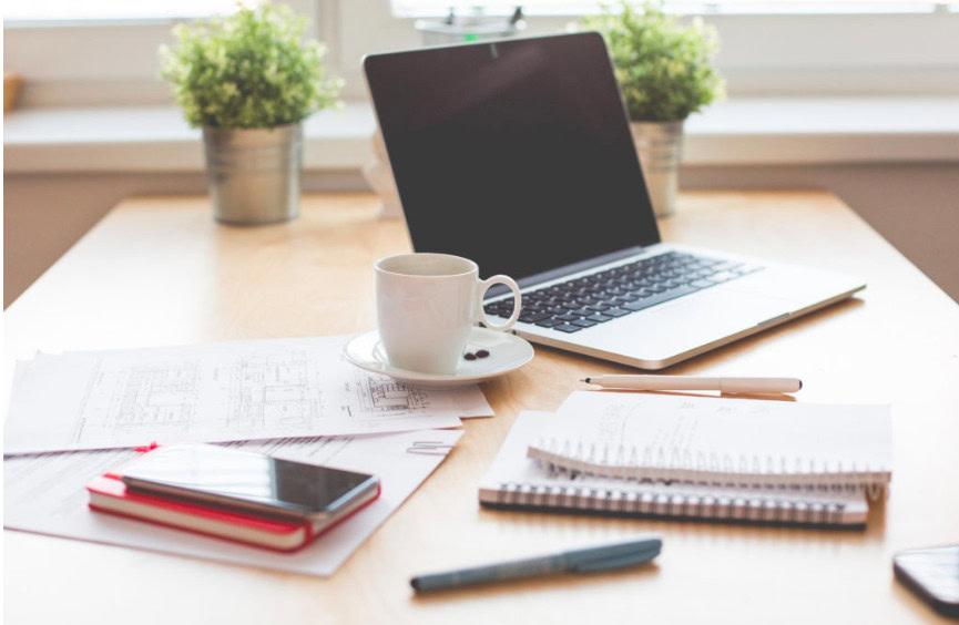 Ein Arbeitsplatz mit Laptop, Handy, Notizbüchern, Stift und Kaffeetasse.