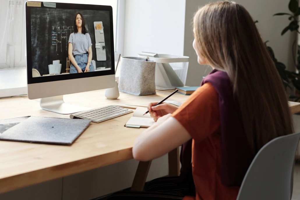 Kind sitzt mit Noitzblock am PC und schaut ein Video von einer Frau in der Schule