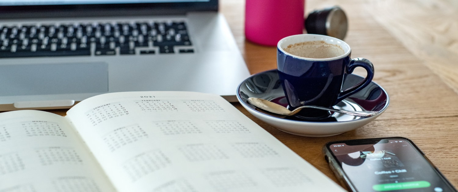 Ein Kalender, ein Smartphone, ein Laptop und eine Tasse Kaffee stehen auf einem Holztisch.