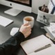 Eine Person hält eine Tasse auf einem Schreibtisch, auf dem Arbeitsmaterialen liegen.
