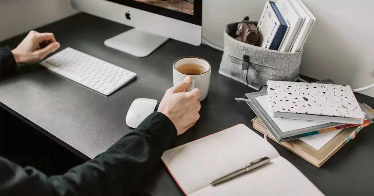 Eine Person hält eine Tasse auf einem Schreibtisch, auf dem Arbeitsmaterialen liegen.