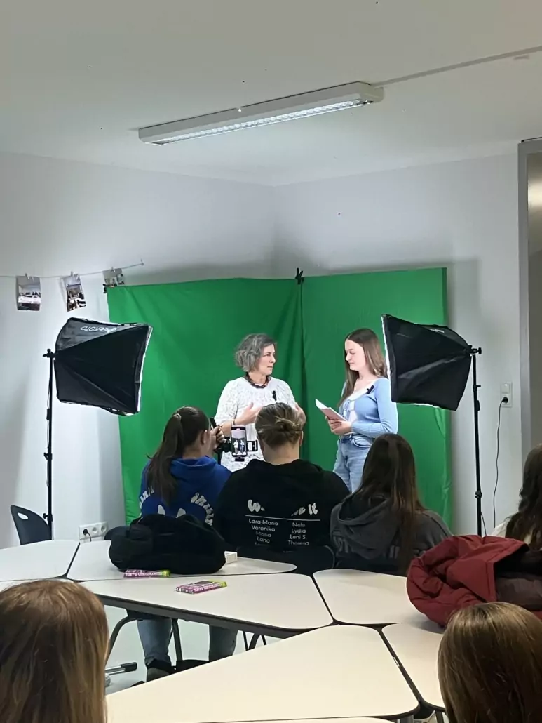 Eine Schülerin interviewt eine Politikerin vor einem Greenscreen. Im Vordergrund sitzen einige Schülerinnen, die zuhören und mitfilmen.