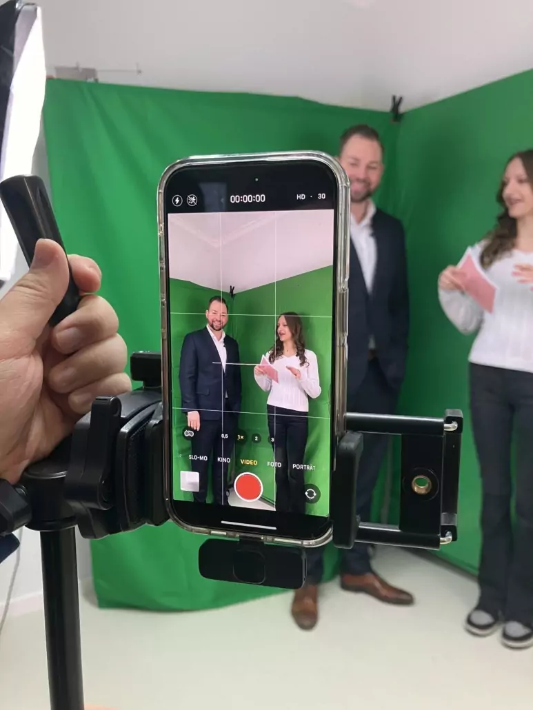 Eine Schülerin interviewt einen Politiker vor einem Greenscreen. Im Vordergrund sieht man ein Handy in einem Stativ, das mitfilmt.