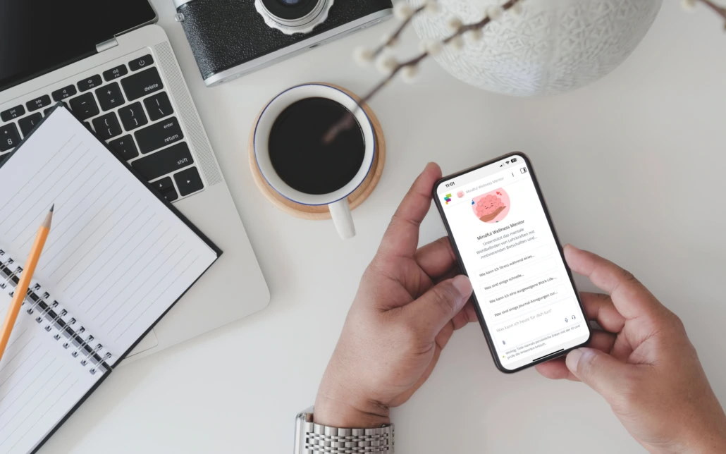 Ansicht von oben: Ein Laptop mit Notizbuch, daneben eine Kafeetasse und eine Vase. Unten rechts halten zwei Hände ein Handy. Auf dem Handy wird der fobizz KI-Assistent „Mindful Wellness Mentor“ angezeigt.